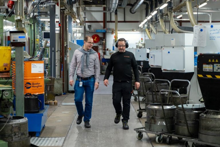 Kaksi miestä kävelee tehdaskäytävää metallintyöstökoneiden vieressä.